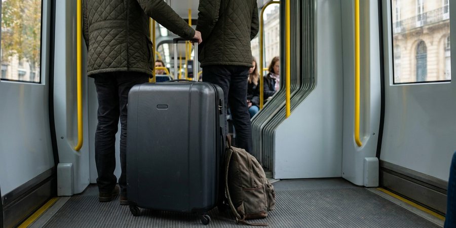 Большой чемодан и рюкзак на полу у двери трамвая, пассажир в стёганой куртке держит ручку чемодана