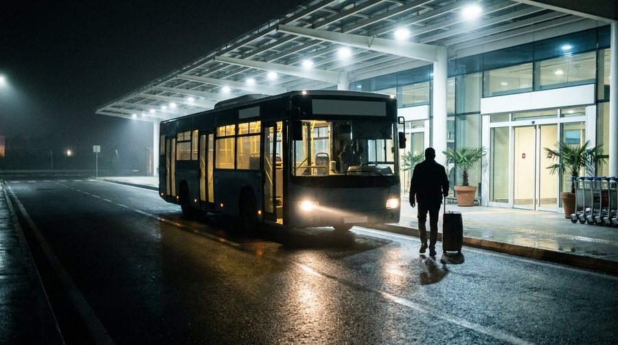 Ночной городской автобус с открытой дверью у стеклянного выхода терминала, к нему идёт пассажир с чемоданом