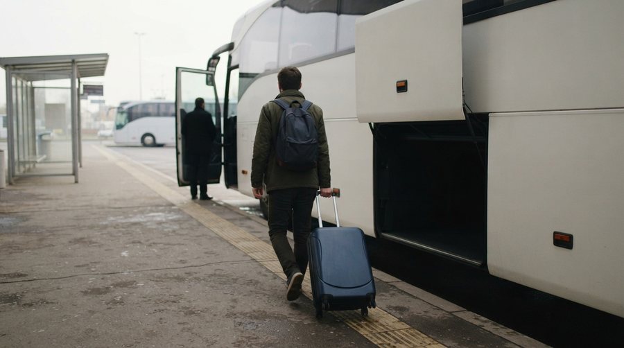 Путешественник катит чемодан к открытому багажному отсеку белого междугороднего автобуса