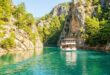 Двухпалубная прогулочная лодка идёт по бирюзовой воде между светлыми скалами, поросшими соснами