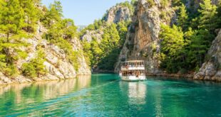 Двухпалубная прогулочная лодка идёт по бирюзовой воде между светлыми скалами, поросшими соснами