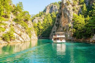 Двухпалубная прогулочная лодка идёт по бирюзовой воде между светлыми скалами, поросшими соснами