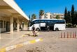 Площадка автовокзала Кушадасы: белый автобус, пассажиры с багажом, навес и кипарисы