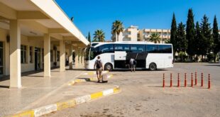 Площадка автовокзала Кушадасы: белый автобус, пассажиры с багажом, навес и кипарисы